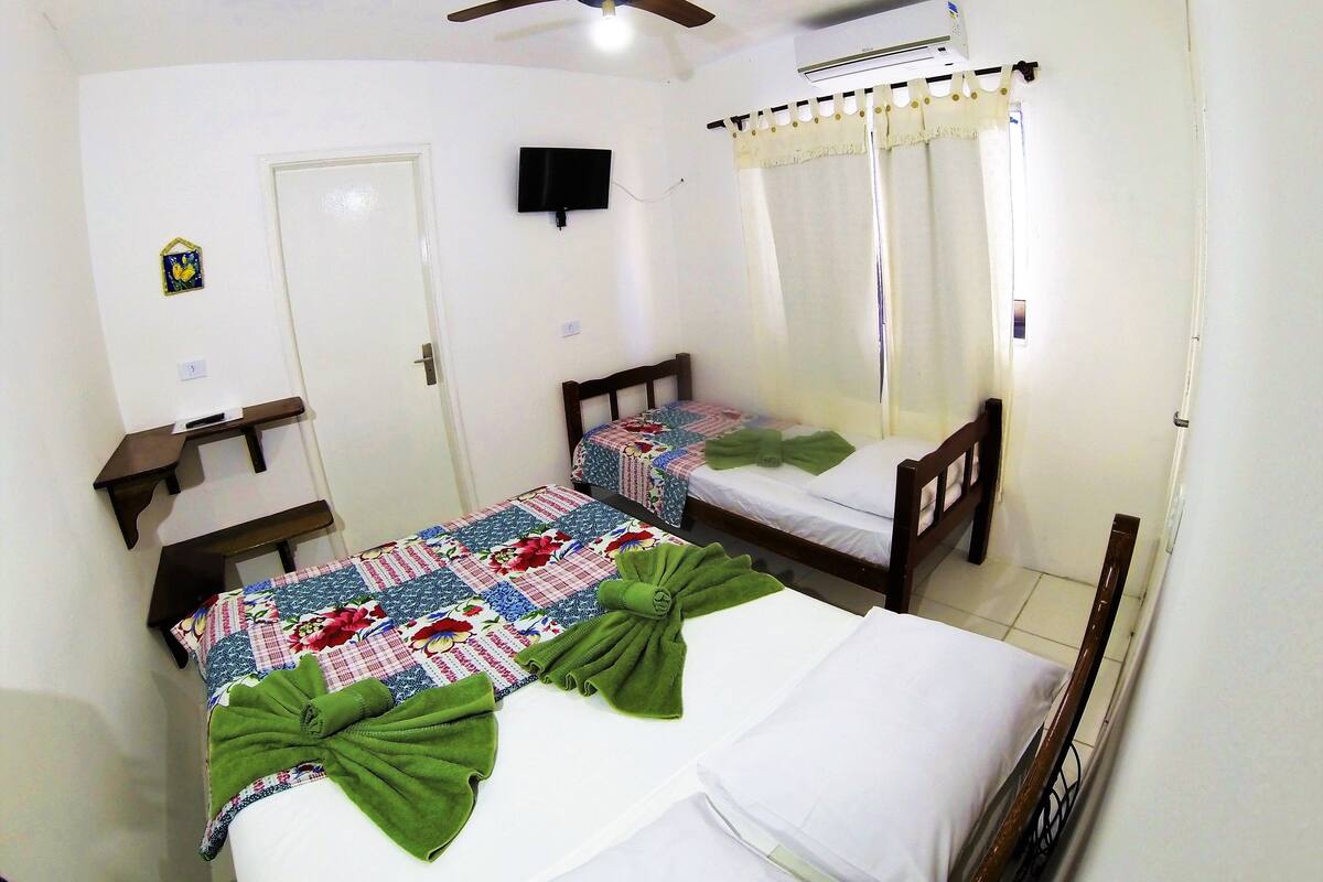 A bedroom with two single beds, a ceiling fan, an air conditioning unit, a TV mounted on the wall, and a small shelf.