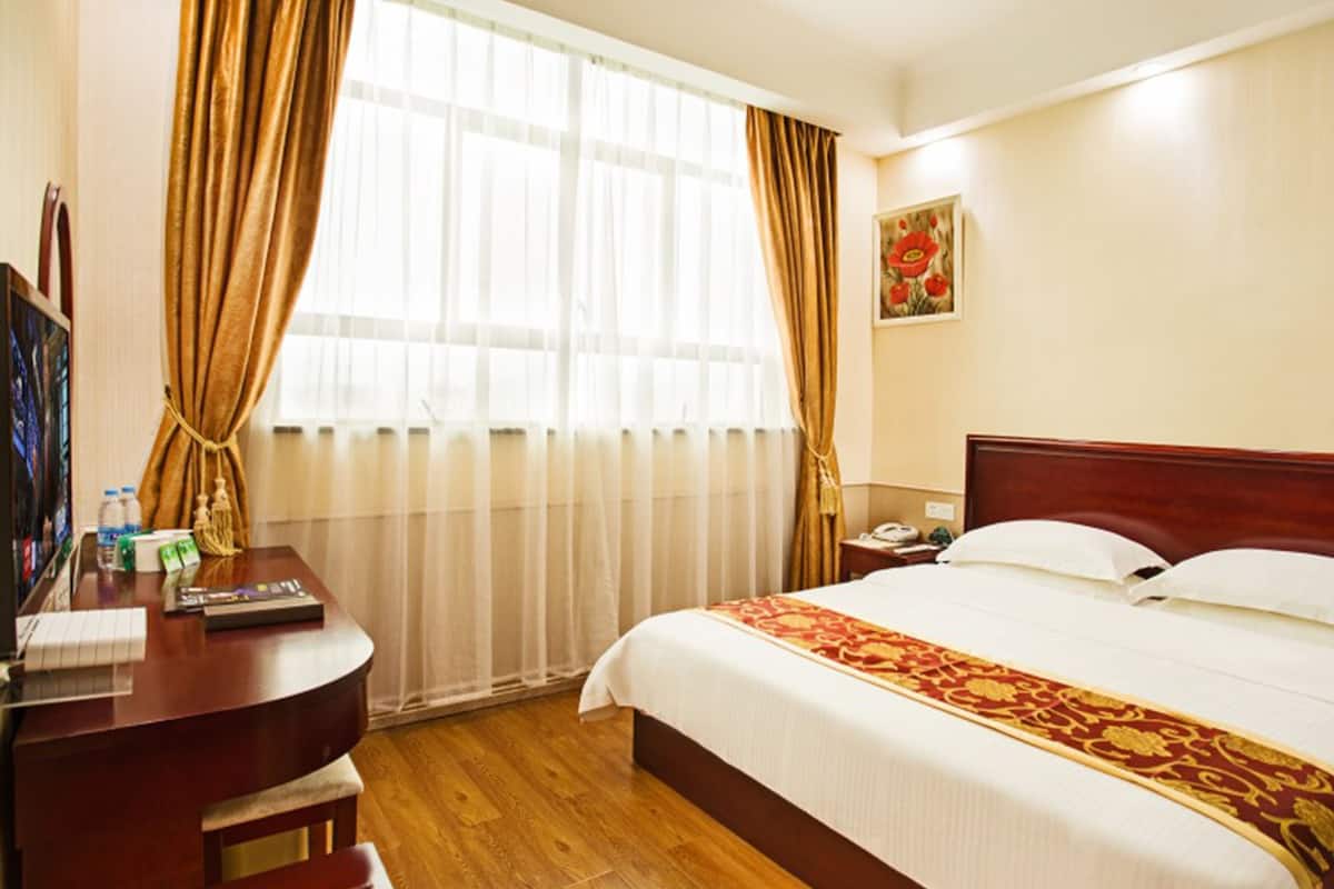 Quarto de hotel com escrivaninha de madeira, cama com colcha vermelha e dourada, mesa de cabeceira com telefone e janela com cortinas leves.