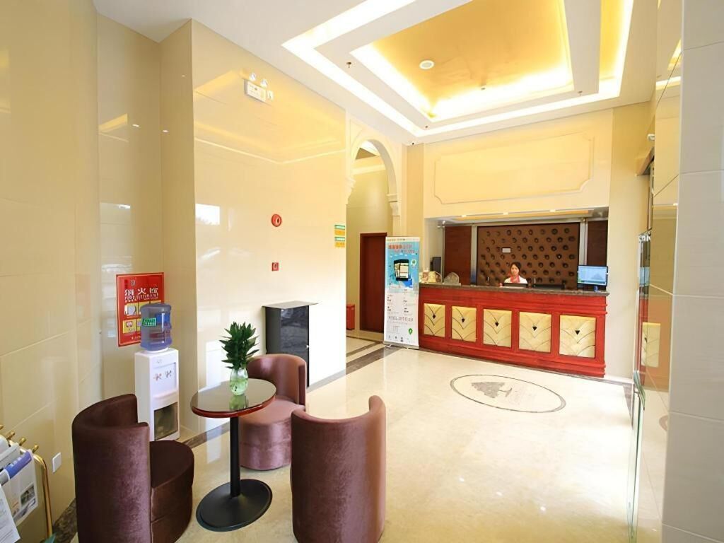 Un vestíbulo de hotel con mostrador de recepción, una pequeña zona de estar con sillas, dispensador de agua y un cartel de seguridad contra incendios.