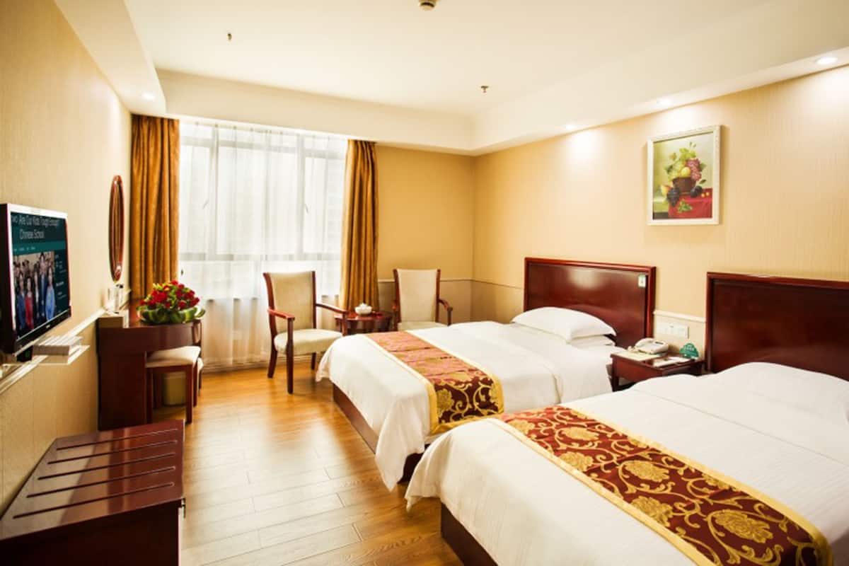 Quarto de hotel com duas camas, uma mesa, uma cadeira, uma televisão e uma janela com cortinas.