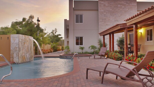 A swimming pool with a stone wall, lounge chairs, and a covered patio area with outdoor seating. - Costa Esmeralda Village (Noord)