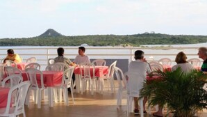 Une terrasse avec des chaises et des tables en plastique blanc, donnant sur une étendue d’eau et une colline.