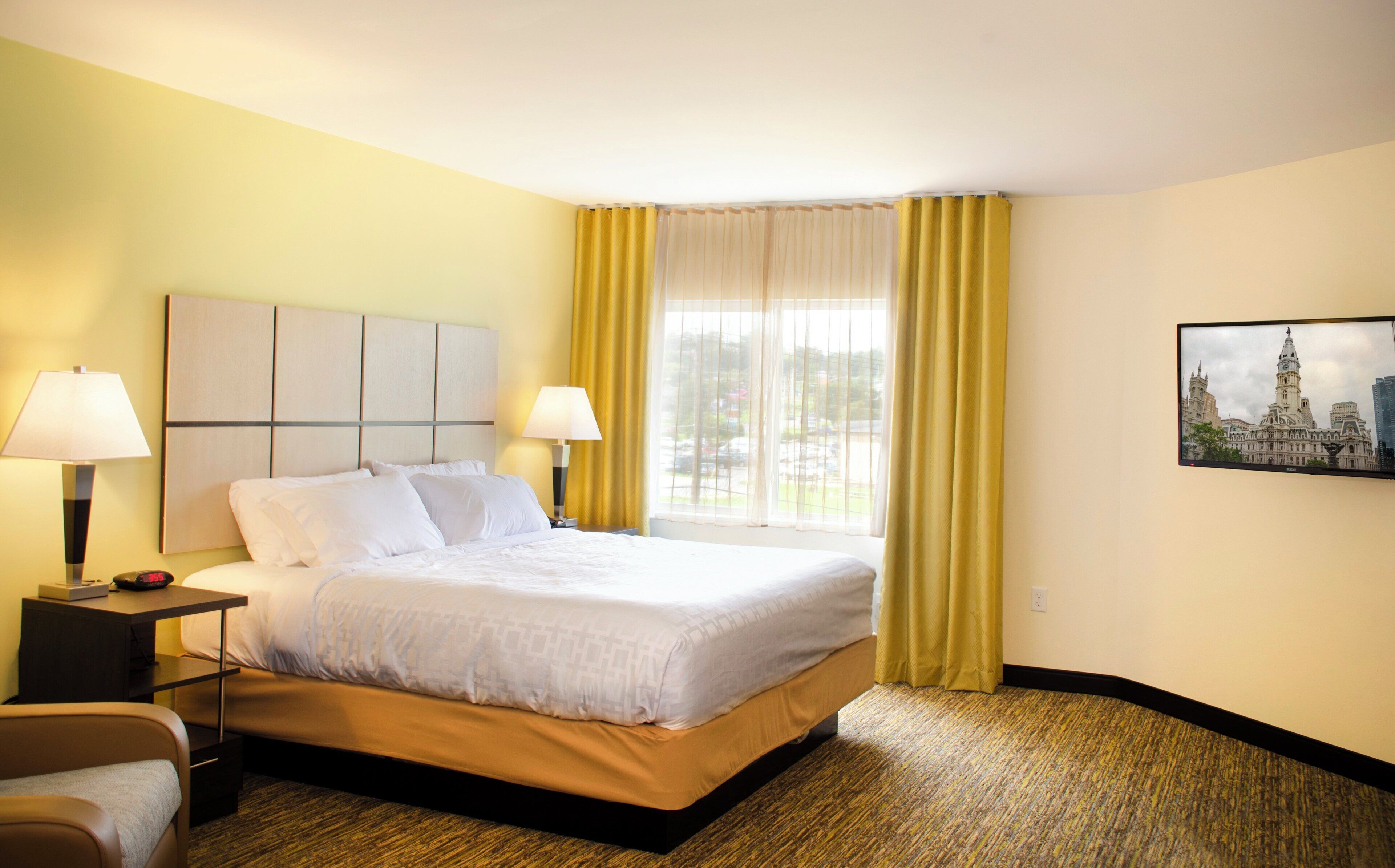 Quarto de hotel com cama, duas luminárias de cabeceira, mesa de apoio, uma cadeira, janela com cortinas amarelas e um quadro na parede.