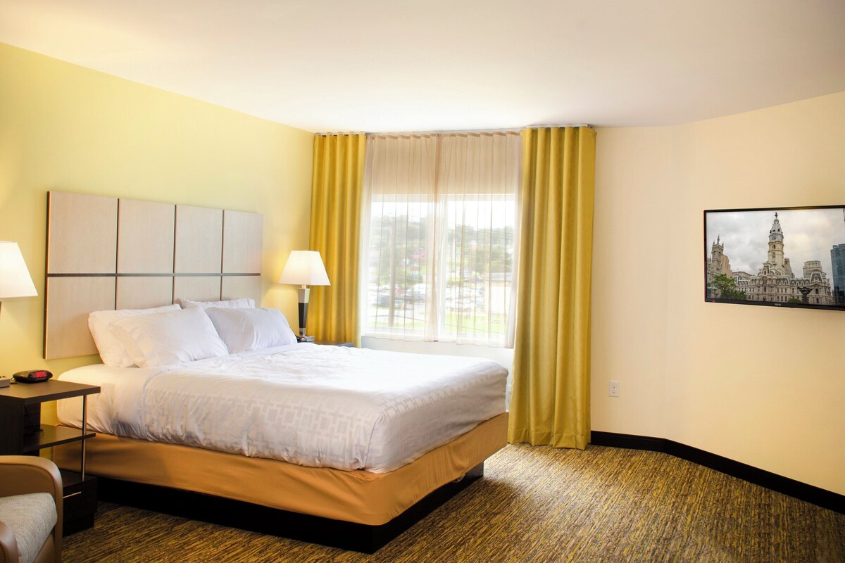 Habitación de hotel con cama, dos lámparas de noche, una mesita de noche, una silla, ventana con cortinas amarillas y un cuadro en la pared.
