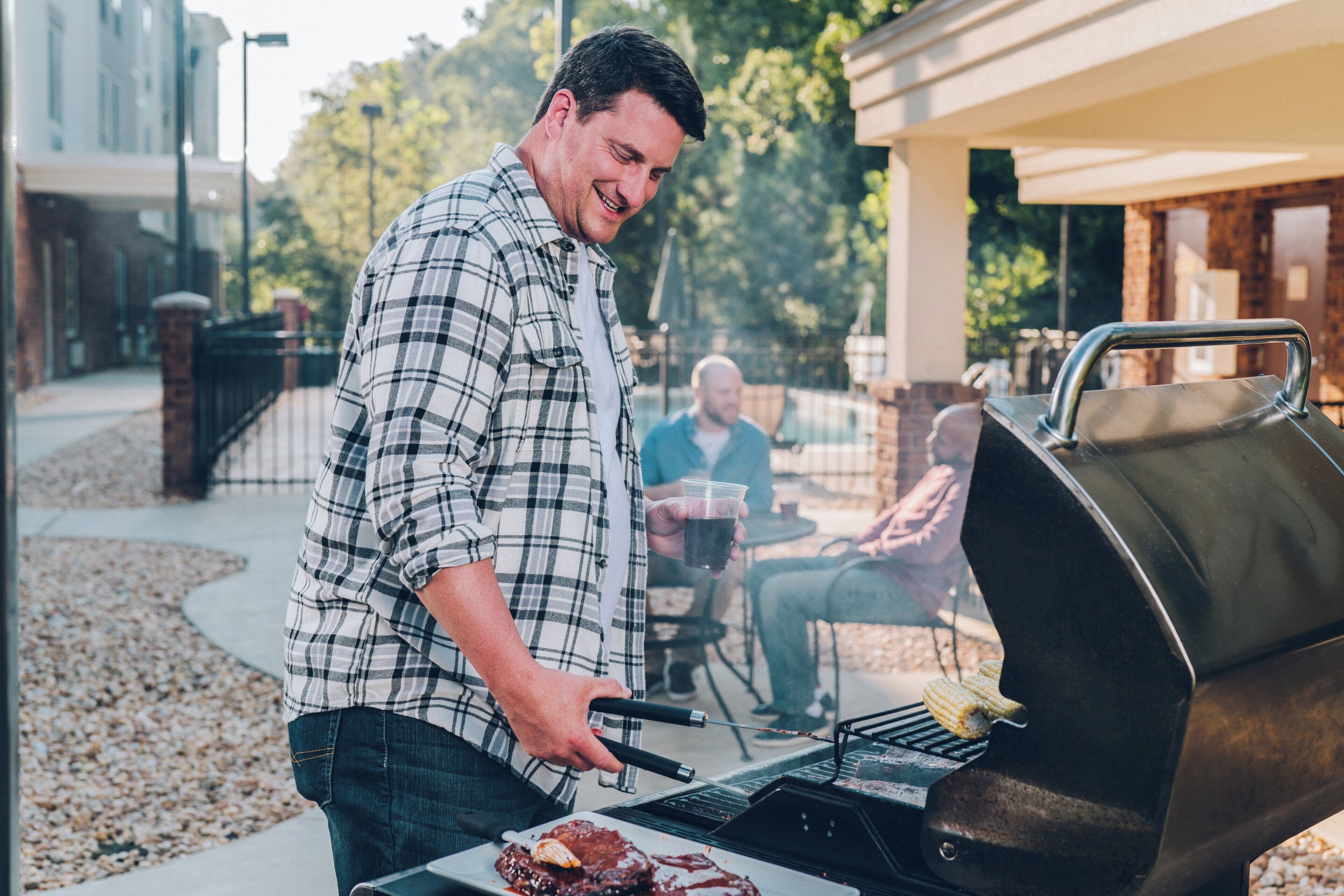 A person grilling meat outdoors with a grill in the foreground and another person sitting in the background.