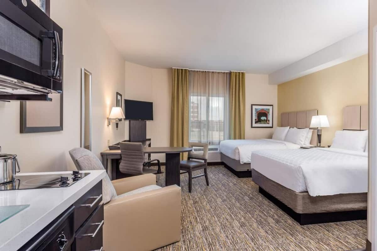 A hotel room with a kitchenette, two beds, a desk with a chair, and a TV.