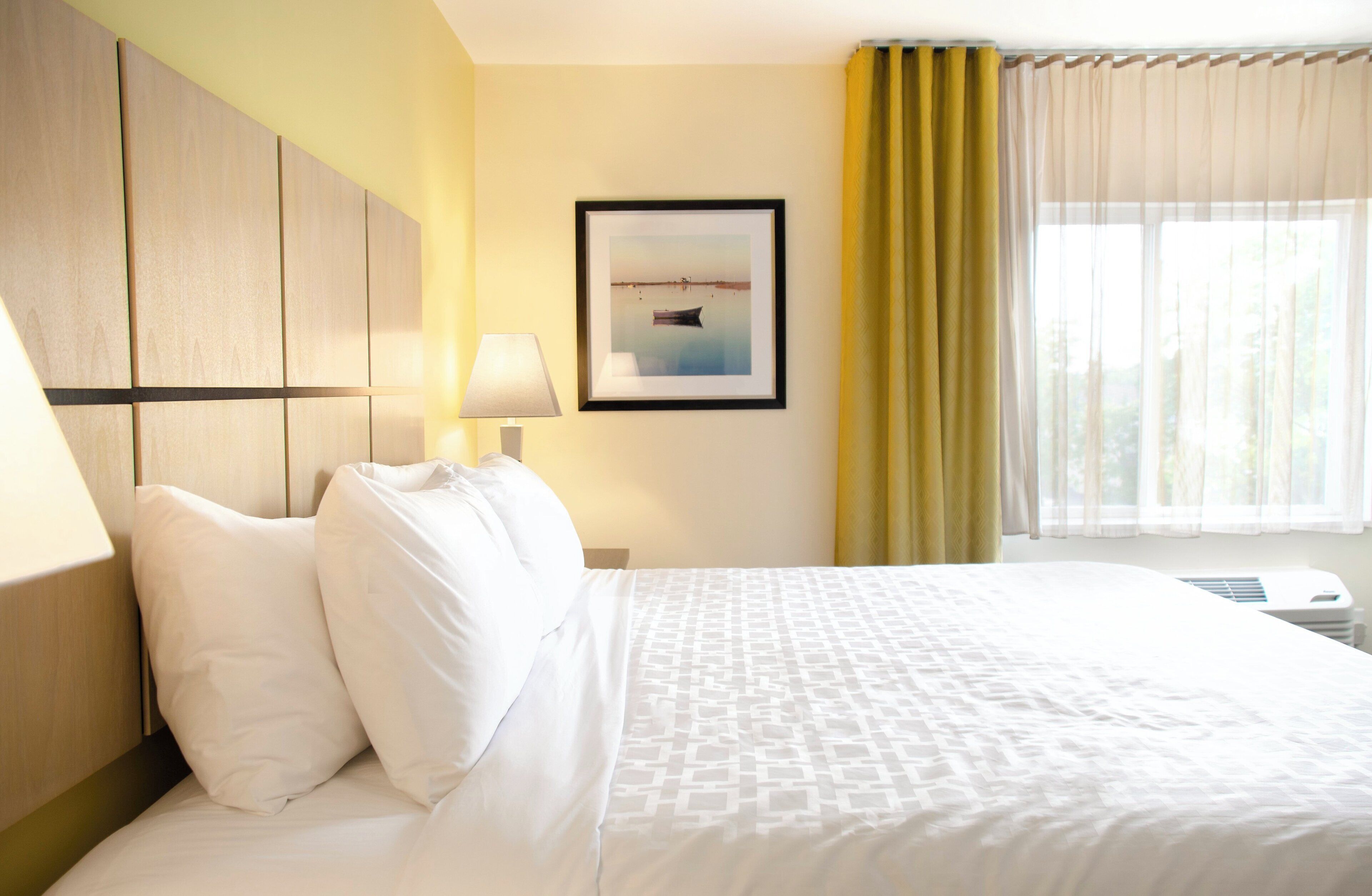 A hotel room with a bed, a nightstand, a lamp, a framed picture, and a window with curtains.