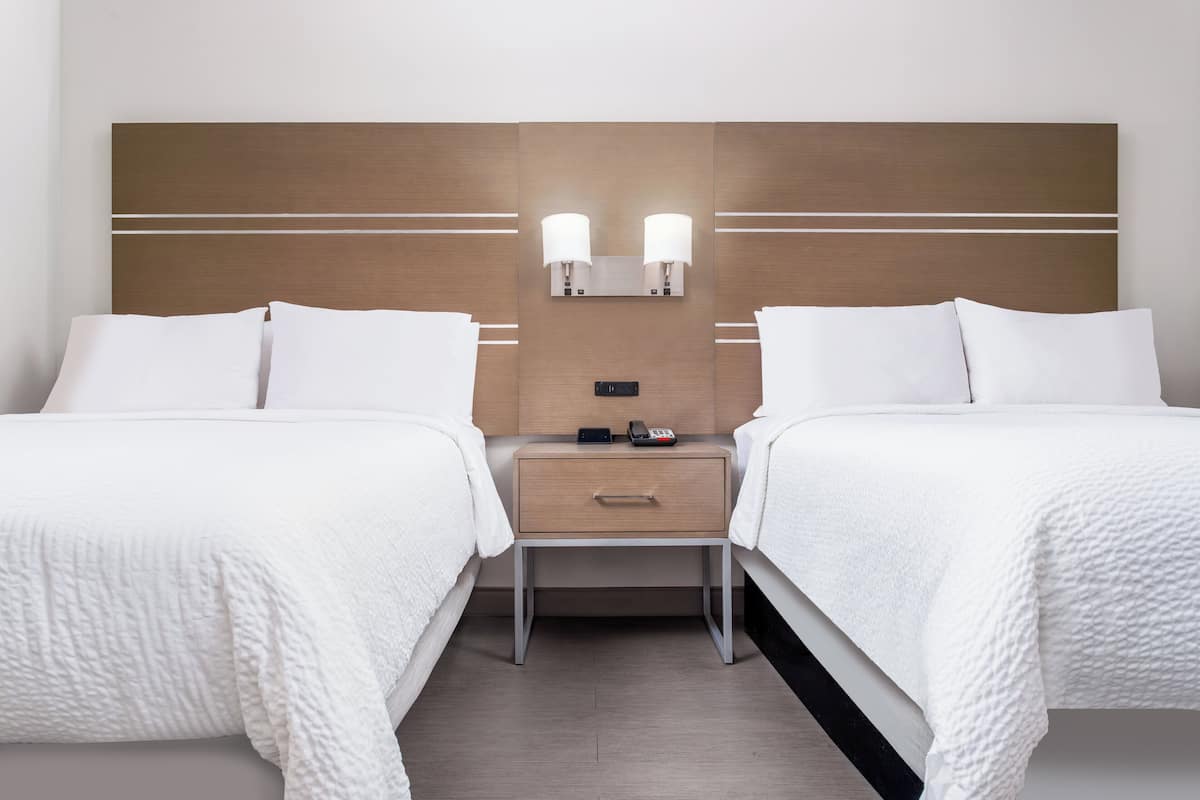 A hotel room with two beds, a nightstand with a lamp, and a control panel.