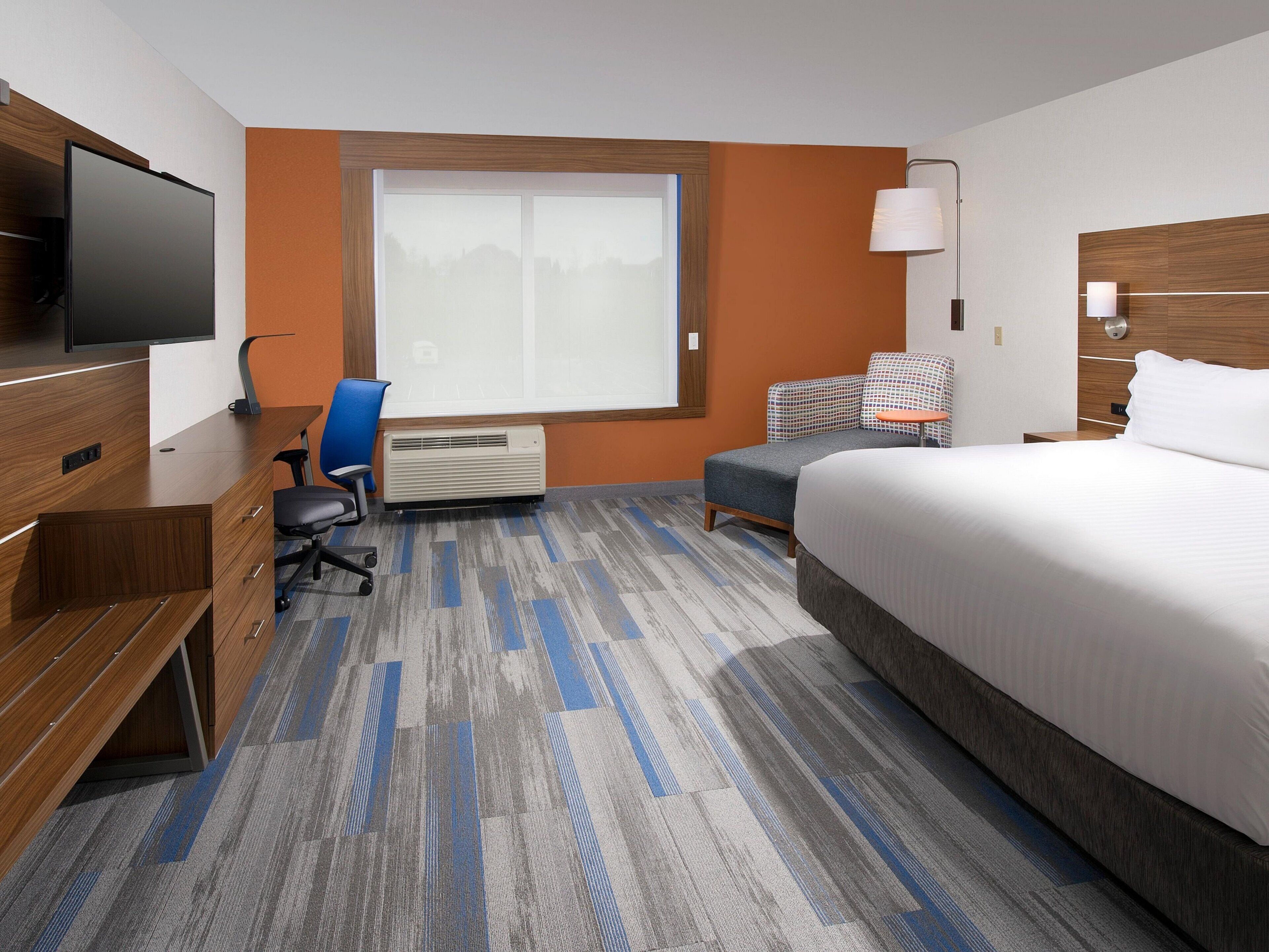 A hotel room with a bed, a desk, a chair, a TV, a window, and a heater.