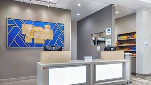 Zona de recepción de hotel con diseño moderno, que incluye un mostrador de recepción, un panel decorativo azul y dorado, y un bar de aperitivos con diversos productos envasados.