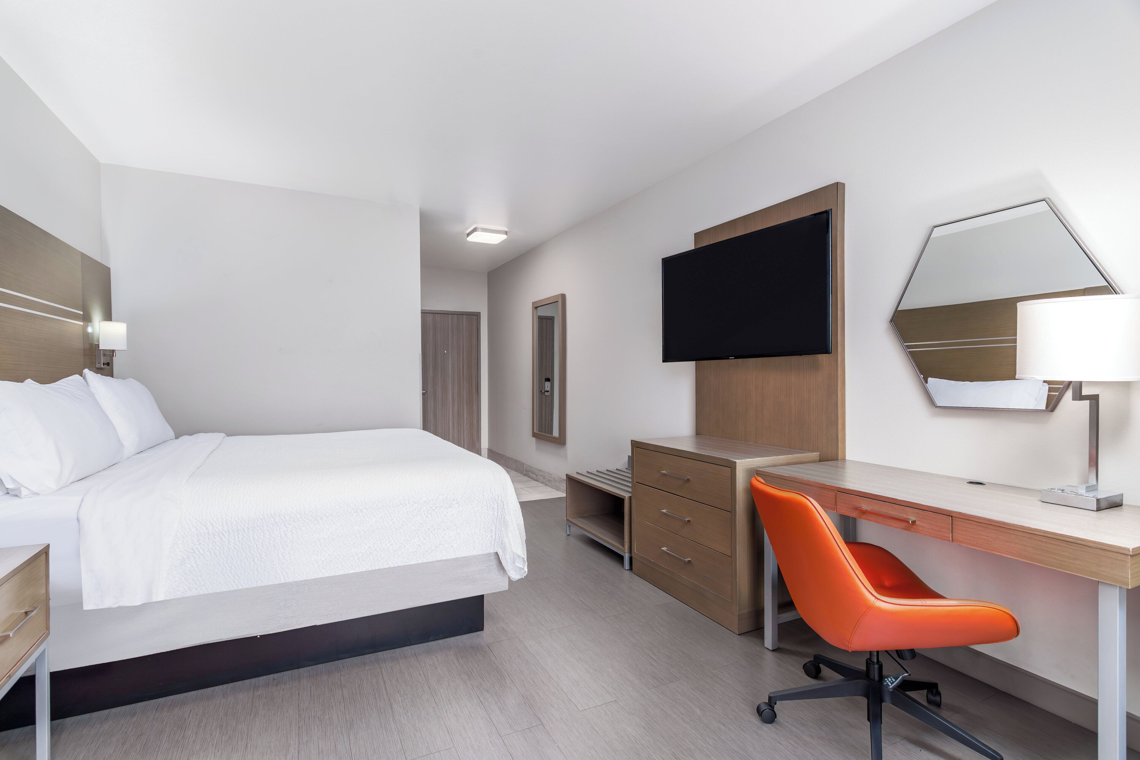 Een moderne hotelkamer met een bed, een bureau met stoel, een flatscreen televisie, een spiegel en een lamp.