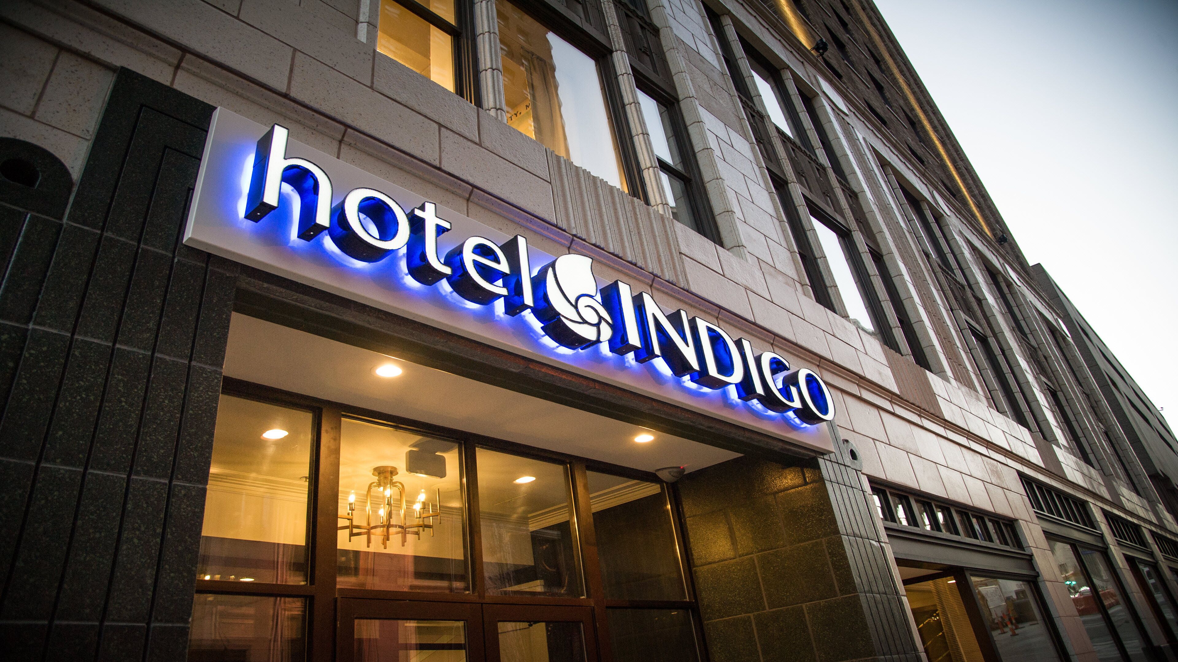 Un letrero de hotel con letras iluminadas, un amplio acceso de vidrio y una fachada de edificio clásica.