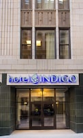 Entrada de hotel con un letrero que dice 'hotel INDIGO', puertas de cristal grandes y el reflejo de un coche en el ventanal.