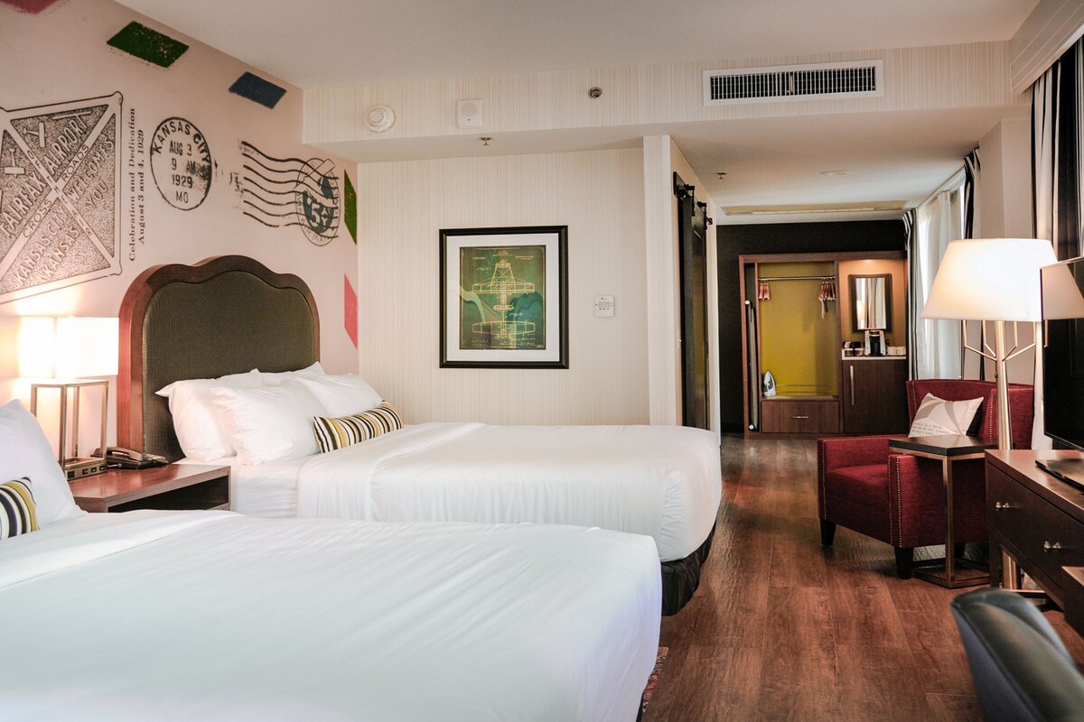 Ein Hotelzimmer mit zwei Betten, einem Schreibtisch mit Lampe, einem Fernseher und einem gerahmten Bild an der Wand.