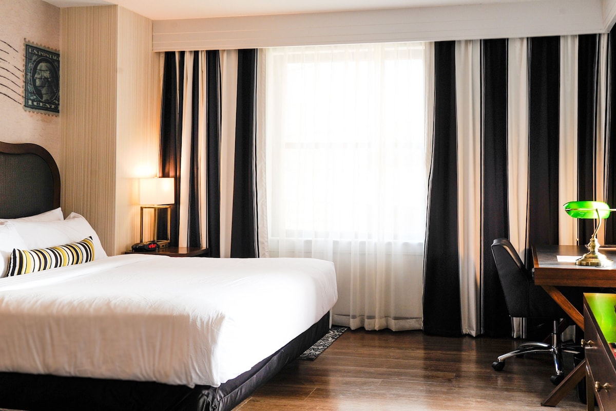 Hotellihuone, jossa on sänky, työpöytä tuolilla, lamppu ja suuri ikkuna, jossa on verhot.
