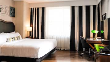 A hotel room with a bed, a desk with a chair, a lamp, and a large window with curtains.