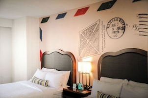 Habitación de hotel con dos camas, una mesita de noche con lámpara, y una pared con un cartel de bienvenida y un sello.