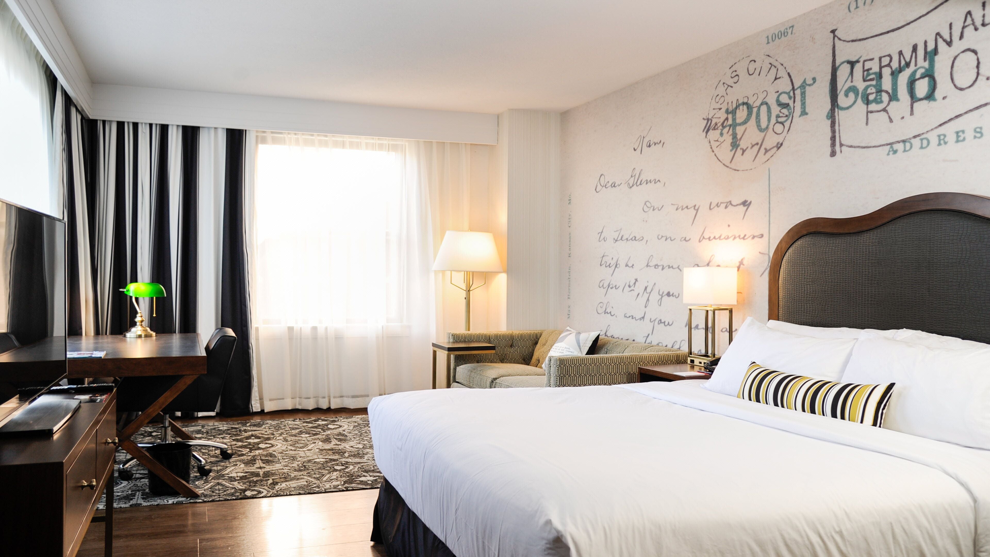 A hotel room with a large bed, a desk, a chair, a lamp, and a wall with handwritten notes.