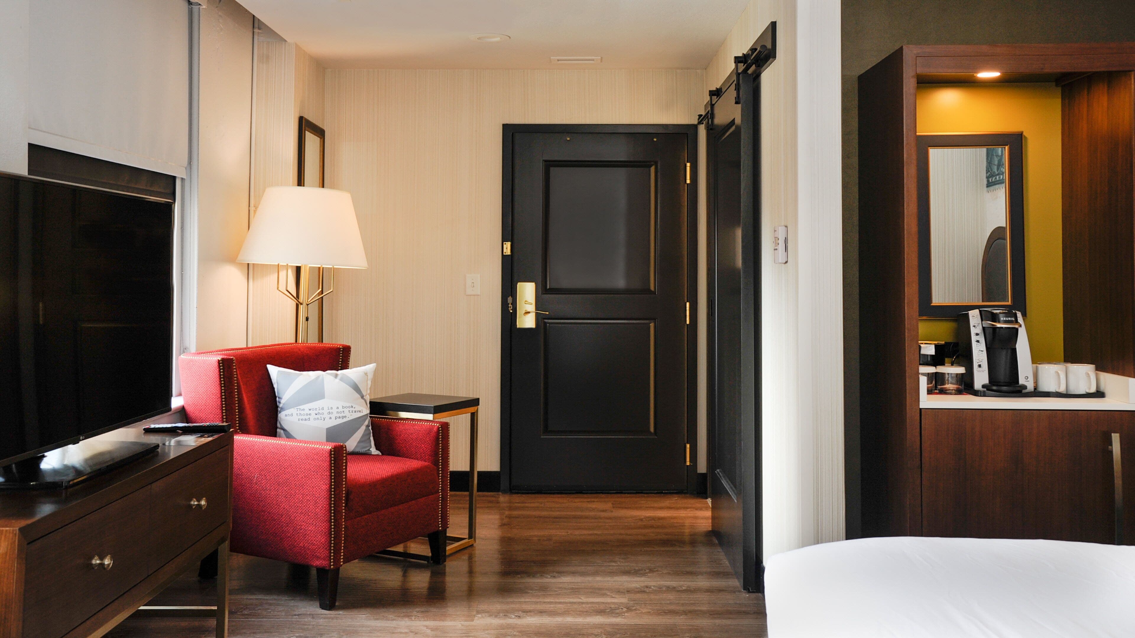 Une chambre d’hôtel équipée d’une télévision, d’un fauteuil rouge, d’une table de chevet avec une lampe, d’une porte noire et d’une machine à café.
