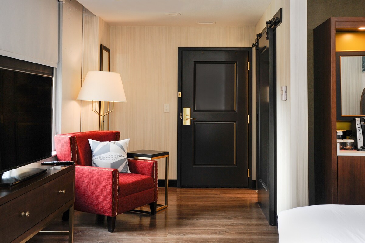 Ein Hotelzimmer mit Fernseher, einem roten Sessel, einem Nachttisch mit Lampe, einer schwarzen Tür und einer Kaffeemaschine.