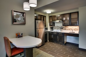 Una cocina moderna con un refrigerador de acero inoxidable, armarios de madera oscura, una encimera blanca y una zona de comedor con una silla y una mesa.