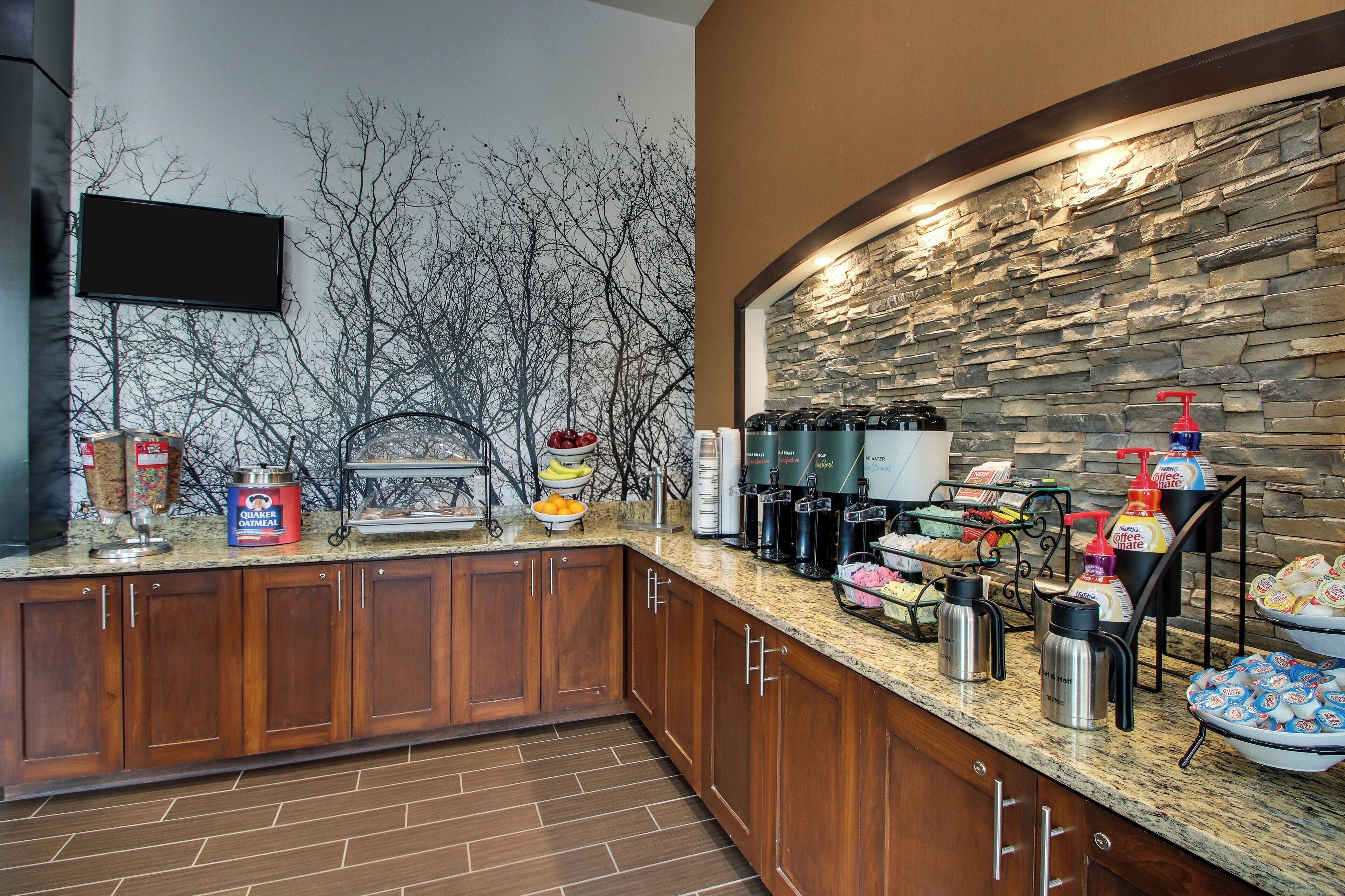 A breakfast bar with a stone wall backdrop, a flat-screen TV, and various food and beverage dispensers.