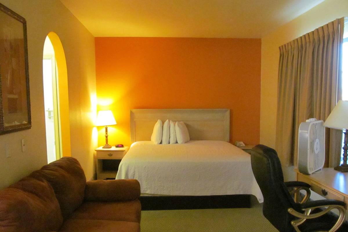 A hotel room with a bed, a nightstand with a lamp, a chair, and a desk with a fan.