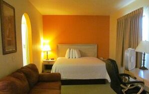 A hotel room with a bed, a nightstand with a lamp, a chair, and a desk with a fan. - Sunbridge Inn & Suites (Zapata)