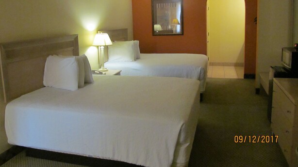 A hotel room with two beds, a nightstand with a lamp, a television, and a mirror on the wall. - Sunbridge Inn & Suites (Zapata)