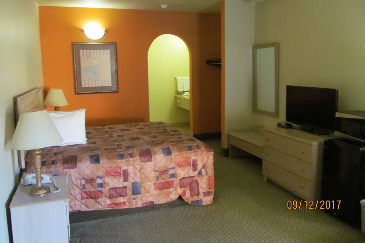 A hotel room with a bed, a desk with a lamp, a dresser, a television, and a framed picture on the wall.