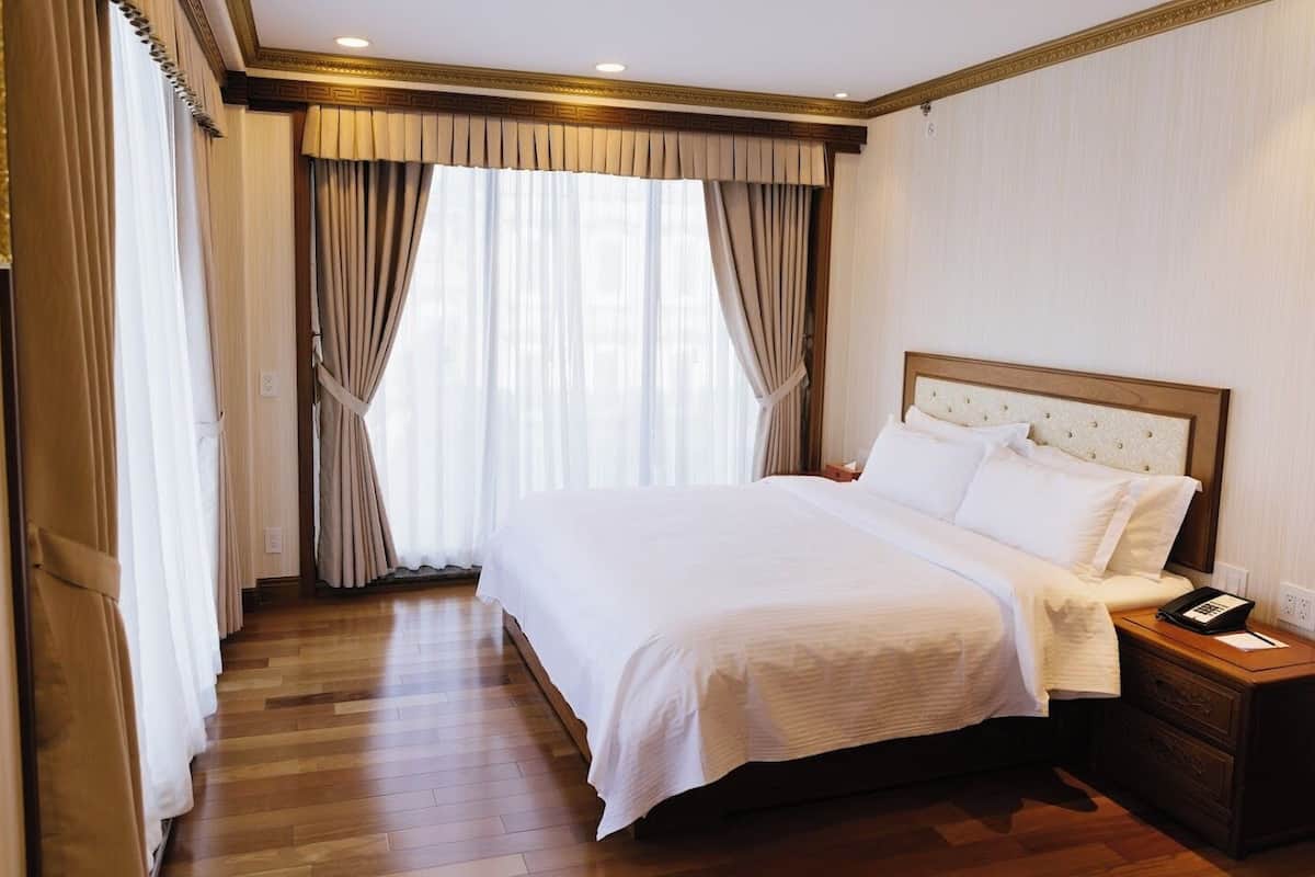 Habitación de hotel con una cama grande, piso de madera, cortinas color beige y una mesita de noche con un teléfono.