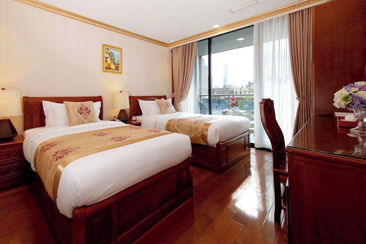 Habitación de hotel con dos camas, muebles de madera, un escritorio con un florero y un ventanal con cortinas.