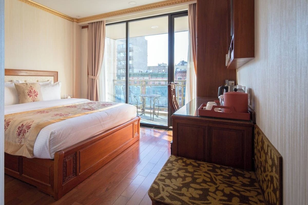 Habitación de hotel con una cama grande, una mesita de noche de madera, una silla y vistas a la ciudad a través de amplios ventanales.