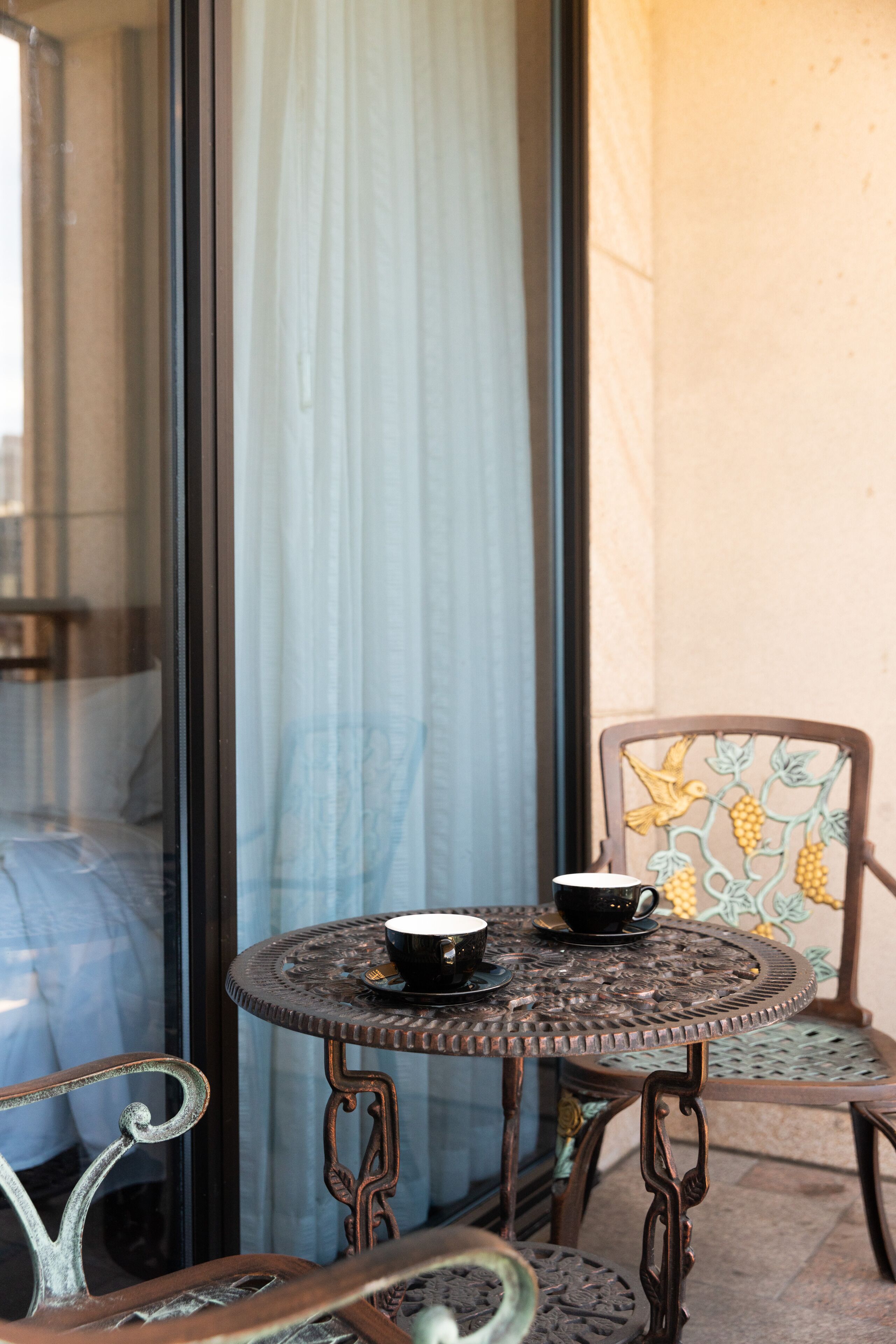 Une table ronde en métal, avec une tasse et une soucoupe, une chaise à motifs floraux et une vue à travers une porte vitrée.