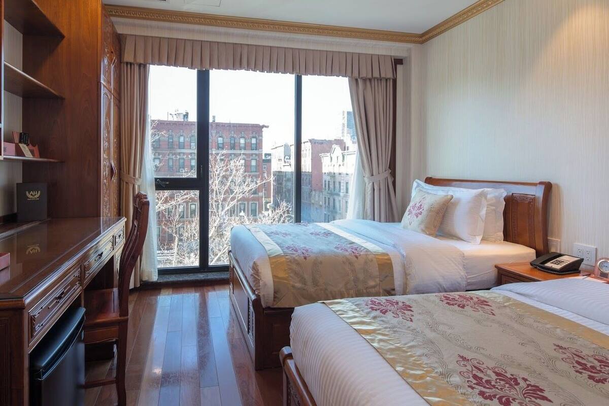 Habitación de hotel con dos camas, un escritorio y una silla. Se aprecia un ventanal con vistas a edificios y árboles.