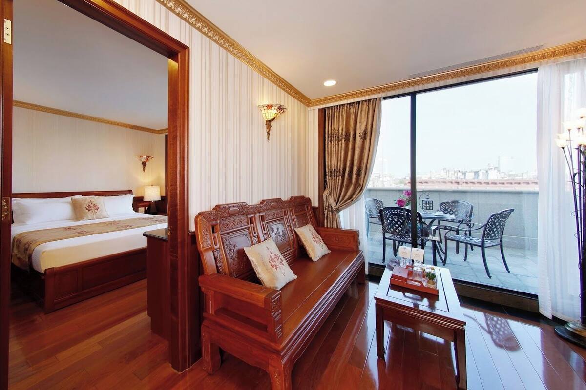 Habitación de hotel con cama, sofá de madera, mesa de comedor y balcón con sillas.