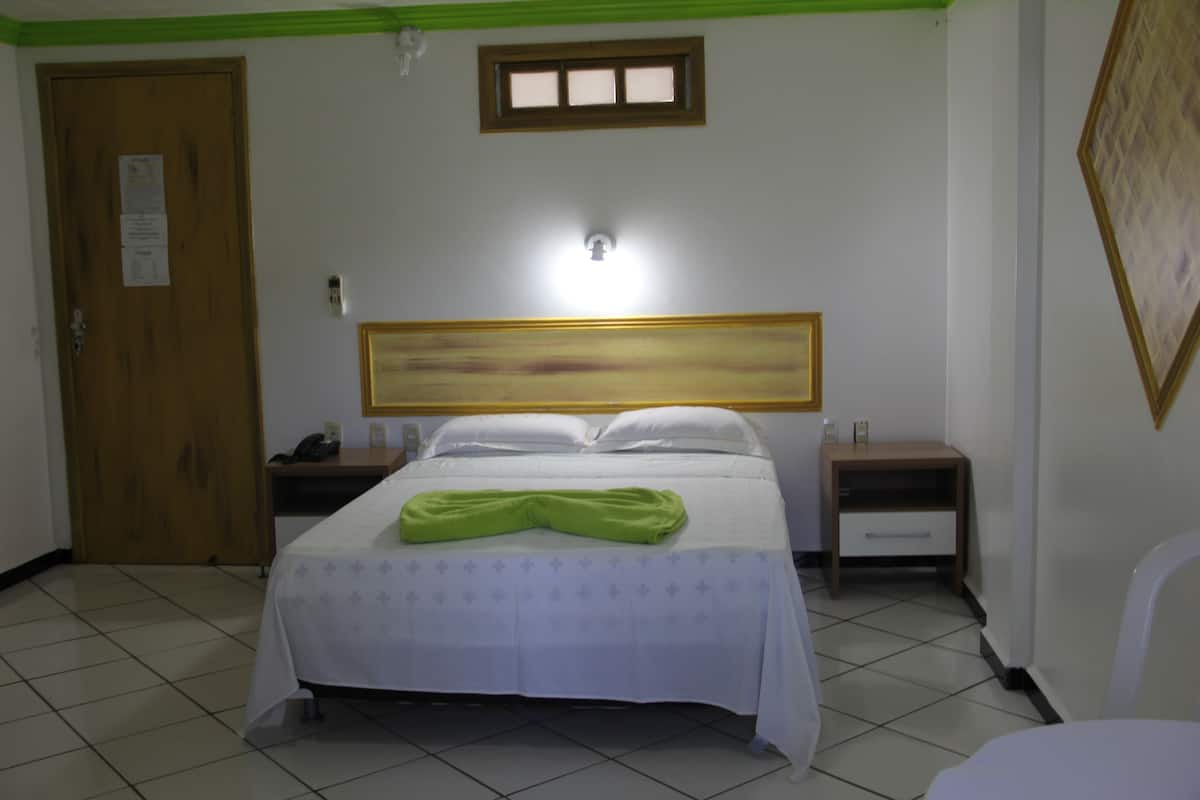 Quarto de hotel com cama, mesas de cabeceira, luminária na parede, porta e uma pequena janela.