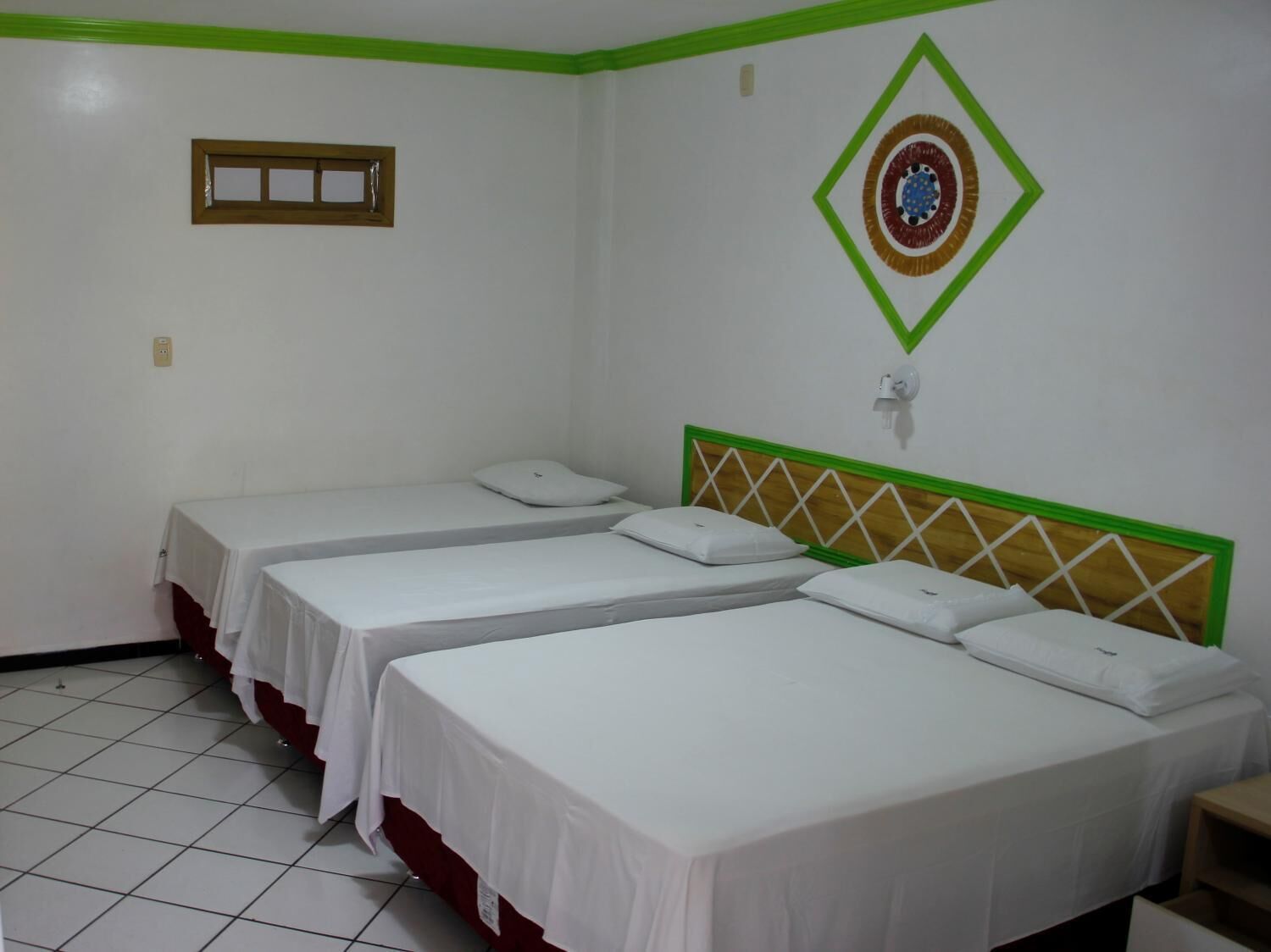 A room with three single beds, each with white bedding and a wooden headboard with green trim. There is a small window on the wall and a decorative diamond-shaped design above the beds.
