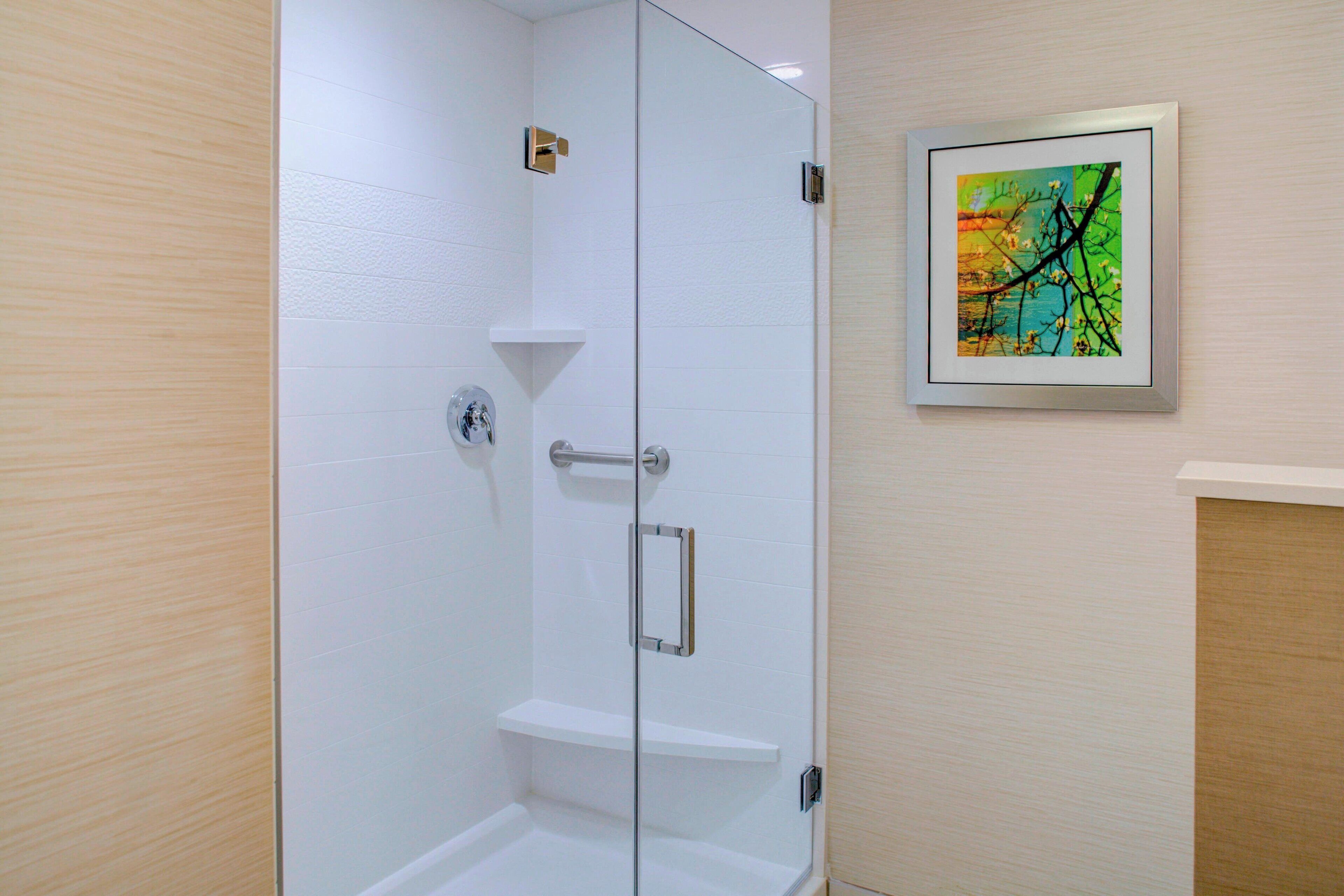 Ducha con cerramiento de vidrio, pared de azulejos blancos, un asa metálica y un estante.