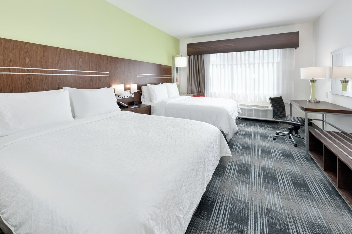 A hotel room with two beds, a desk, a chair, a lamp, and a large window with curtains.
