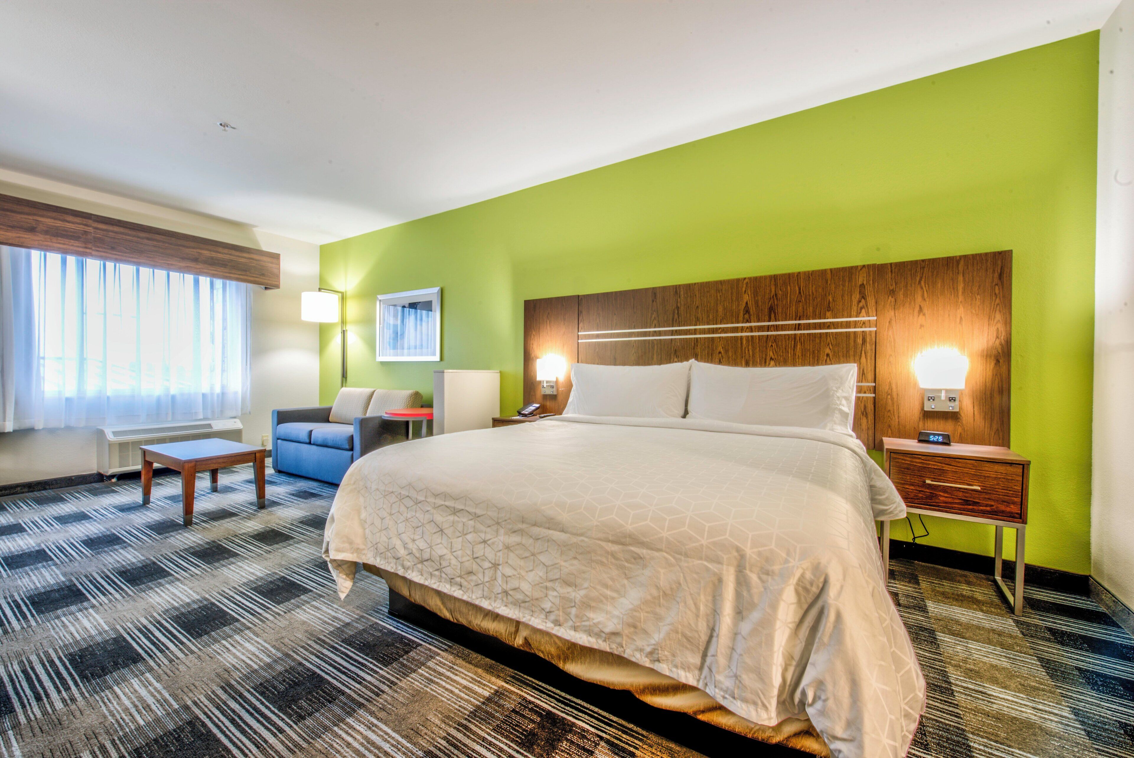 A hotel room with a large bed, a seating area with a chair and a small table, a bedside table with a lamp, and a window with curtains.