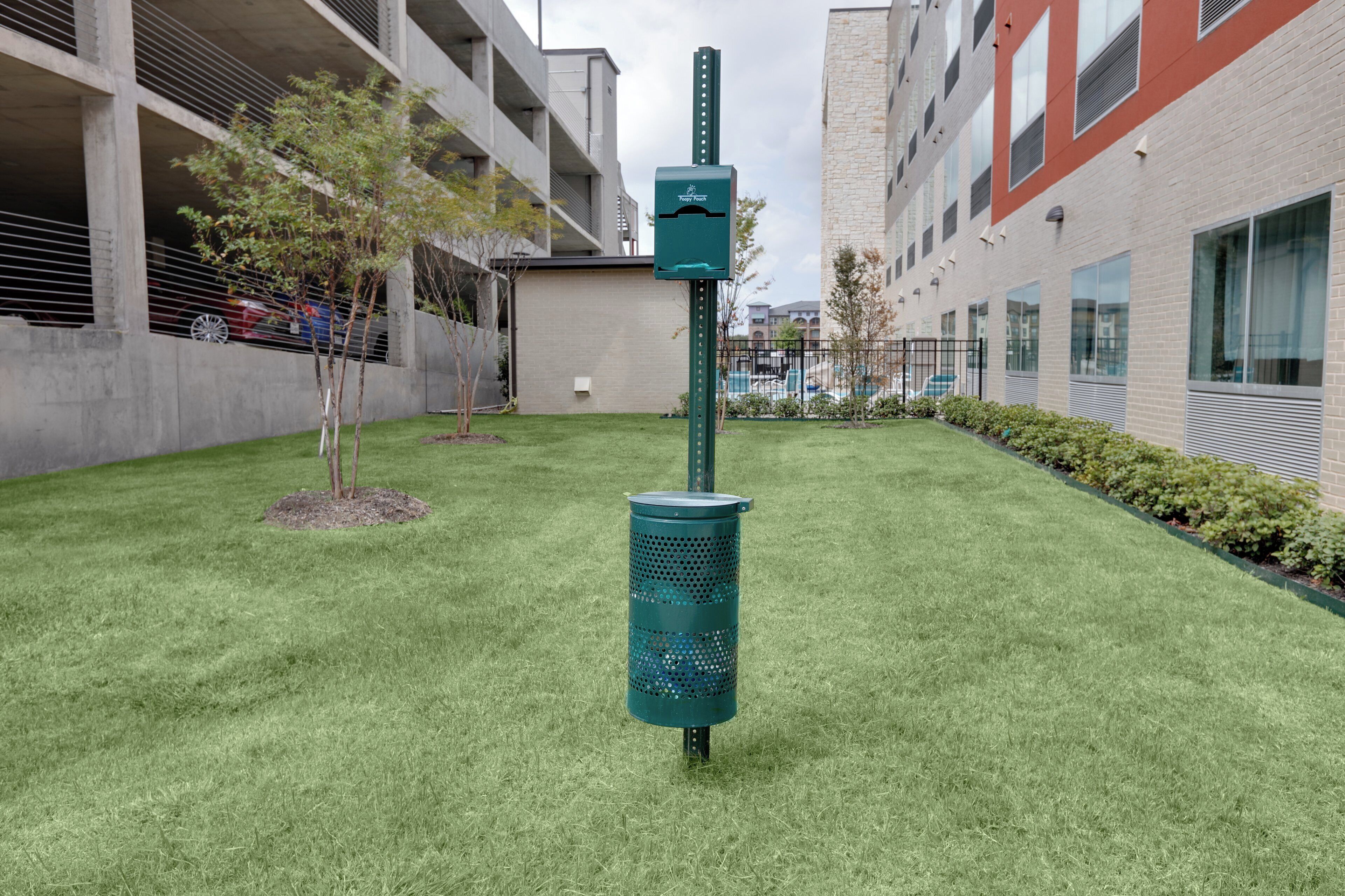 Un bote verde para basura con una etiqueta, un árbol pequeño y un edificio con fachada roja y color crema.