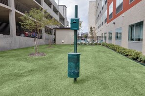 A green trash bin with a label, a small tree, and a building with a red and beige facade. - Holiday Inn Express & Suites Dallas NW - Farmers Branch by IHG (Farmers Branch)