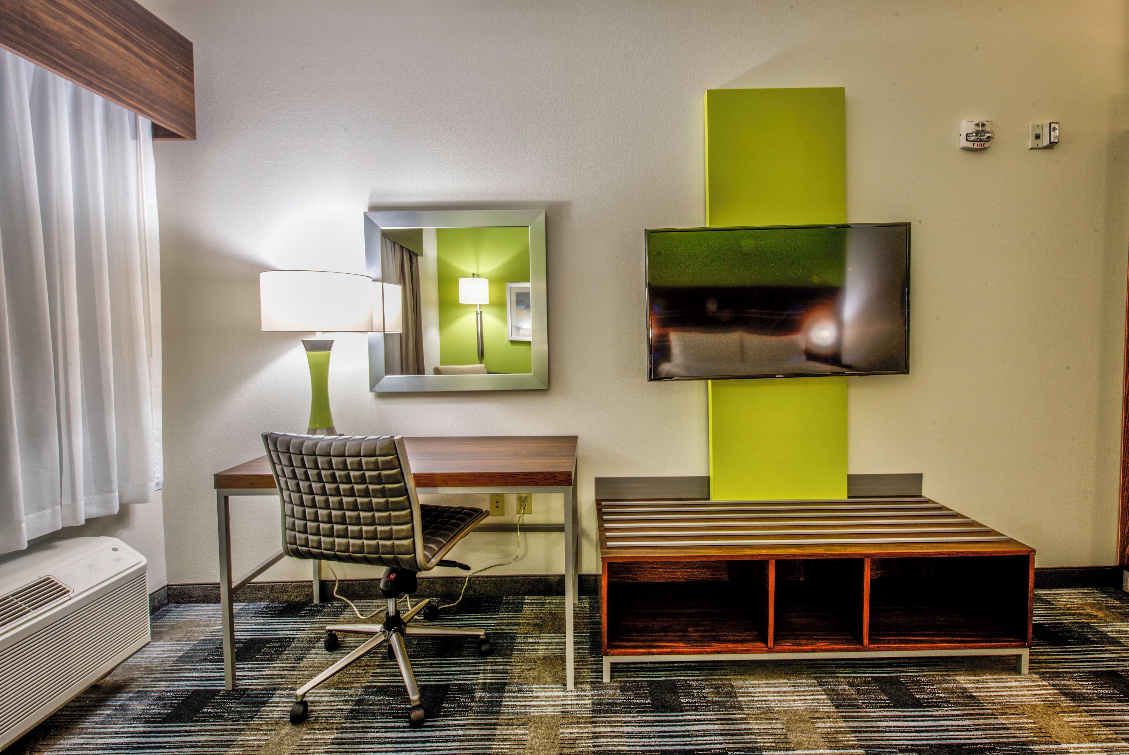 A hotel room with a desk, chair, lamp, TV, and a mirror.