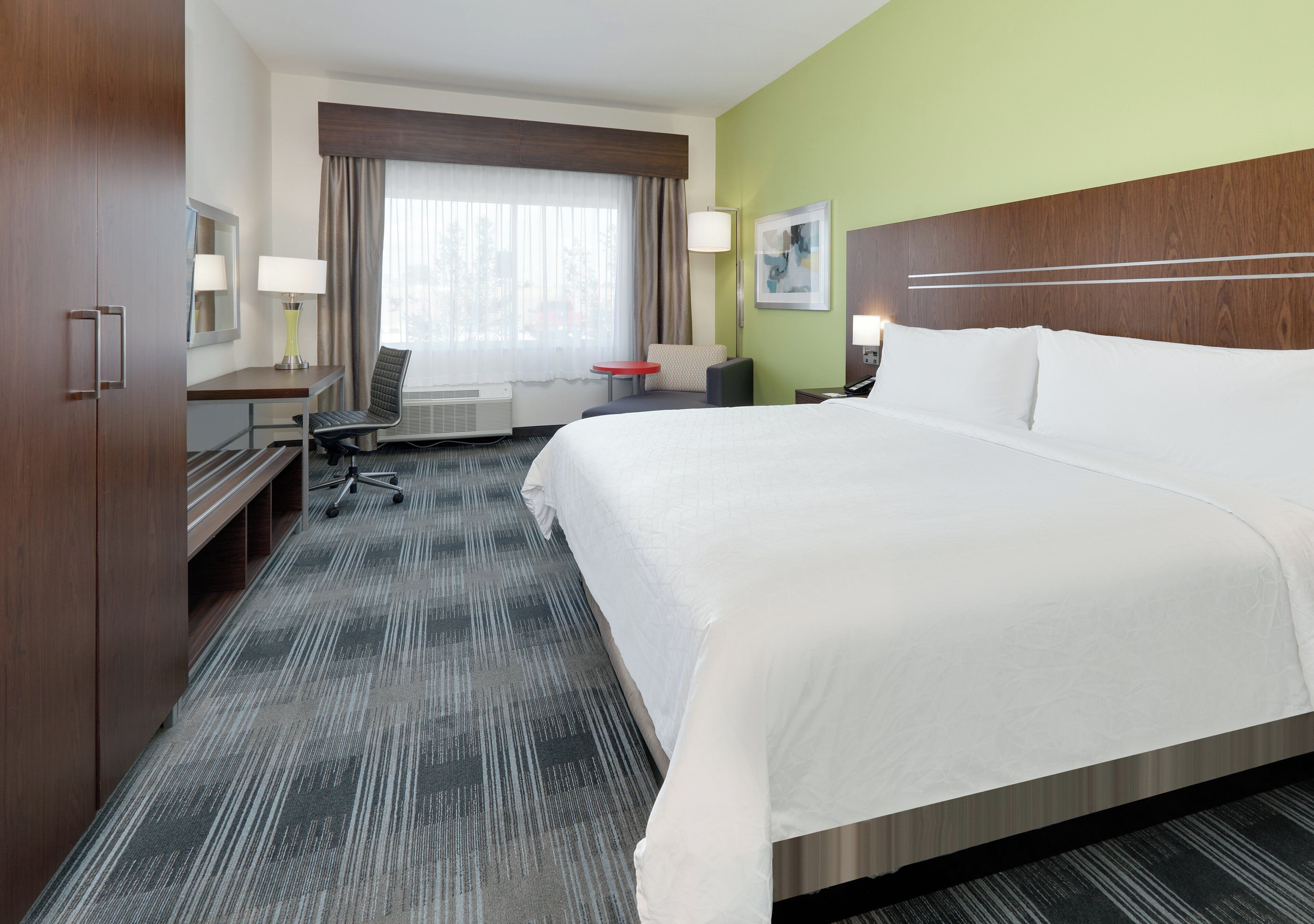 A hotel room with a large bed, a desk with a chair, a small table, a lamp, and a window with curtains.