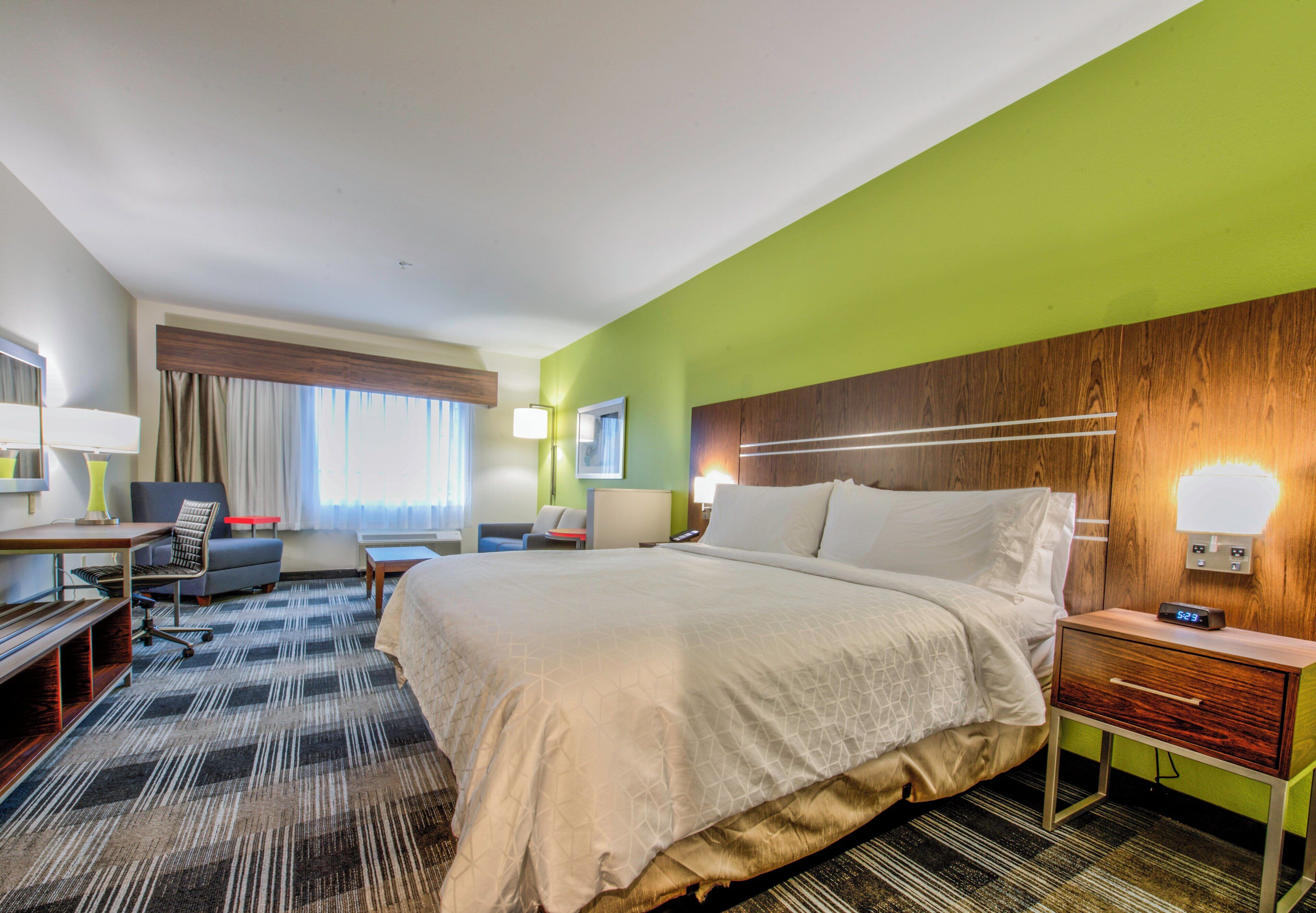 Una habitación de hotel con una cama grande, un escritorio, una silla, una lámpara y una mesita de noche con un reloj.