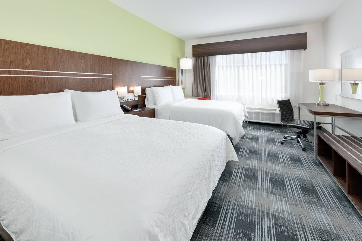 A hotel room with two beds, a desk, a chair, a lamp, and a large window with curtains.