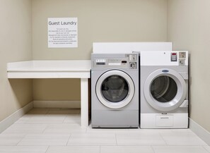 A guest laundry area with two front-loading washing machines, a white counter, and a sign with laundry instructions. - Holiday Inn Express & Suites Dallas NW - Farmers Branch by IHG (Farmers Branch)