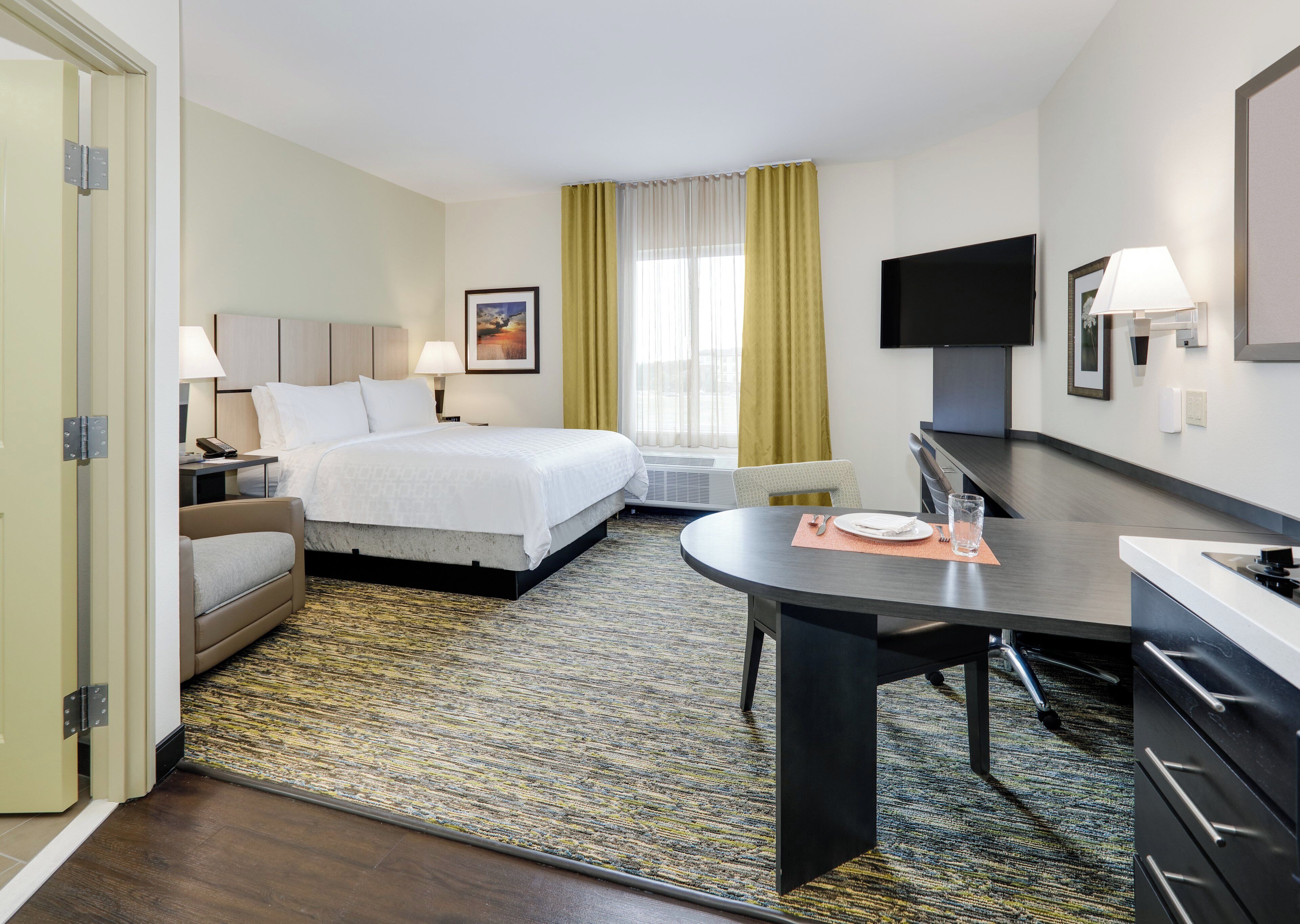 Habitación de hotel con cama, mesita de noche, lámpara, televisor, escritorio, silla y ventana con cortinas.