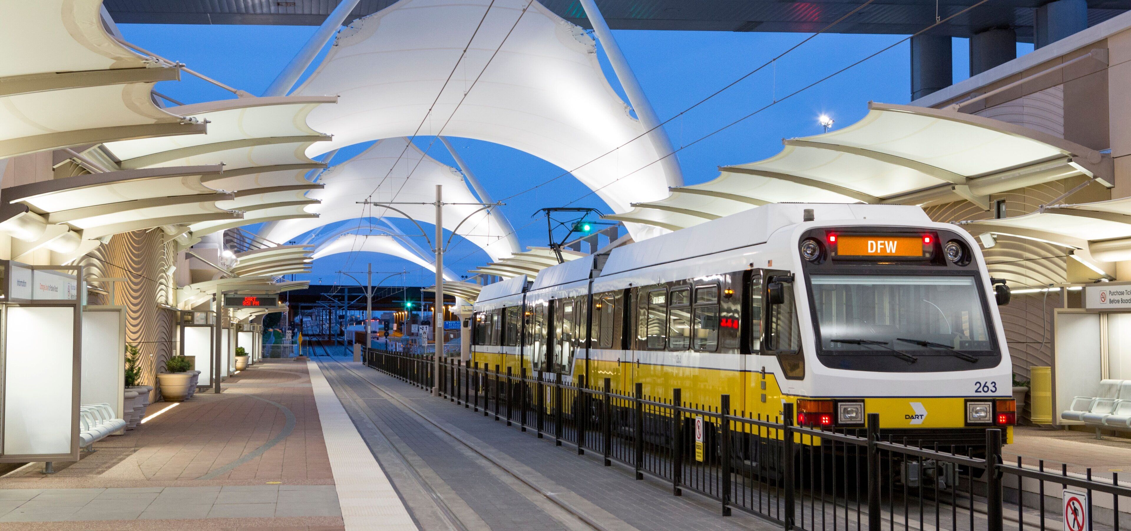 A tram at a station with a modern architectural design, including a curved roof structure and a digital display showing the time.